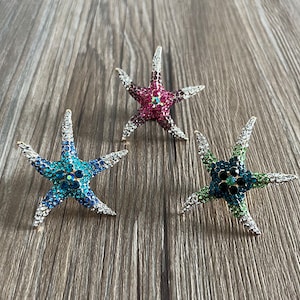 May include: Three jeweled starfish brooches in blue, green, and pink. Each brooch is made of metal and has a star shape with a crystal-like finish.