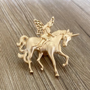 May include: A gold-colored brooch featuring a fairy riding a unicorn. The fairy has detailed wings and is seated on the unicorn's back. The unicorn has a spiraled horn and is in a trotting pose. The brooch is made of metal.