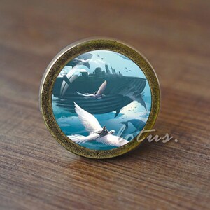 May include: A round, bronze-colored cabinet knob with a blue and white image of a whale and two white doves flying in the sky. The whale is flying over a city skyline.