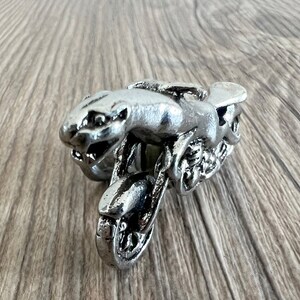 May include: A silver-coloured metal figurine of a motorcycle with a snarling animal figure riding it. The motorcycle has a single wheel and a detailed design.