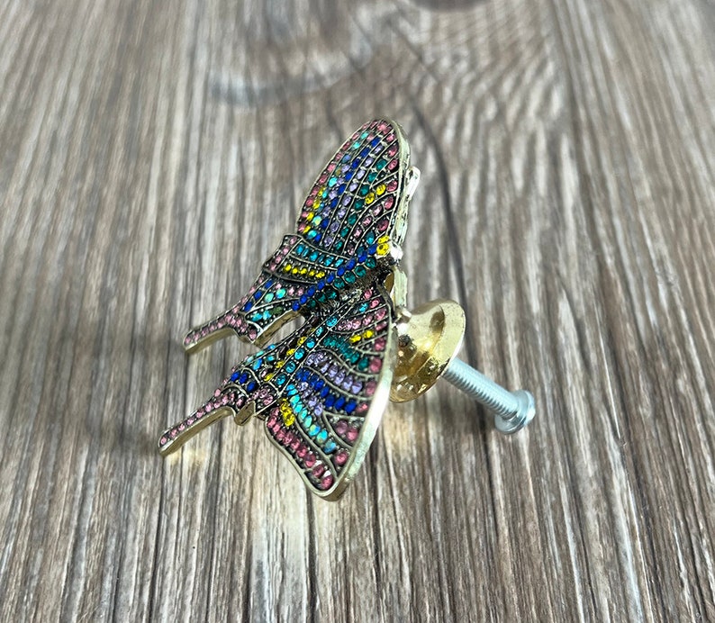 May include: A gold butterfly-shaped cabinet knob with a colourful mosaic design. The knob has a screw for attaching it to a cabinet door.