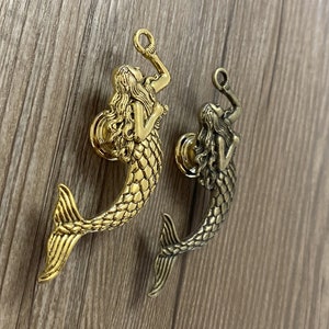 K&ouml;nnte beinhalten: Zwei goldfarbene Meerjungfrau-T&uuml;rklinken mit detailliertem Design. Die Meerjungfrauen halten ihre Schw&auml;nze und haben langes, flie&szlig;endes Haar.
