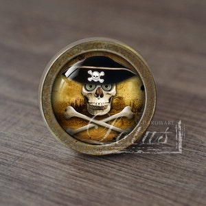 Pode incluir: Um botão de armário redondo de cor latão com um design de caveira e ossos cruzados. A caveira está usando um chapéu de pirata preto com um emblema de caveira e ossos cruzados branco. O fundo é um pergaminho amarelado e desgastado com uma cena de floresta.