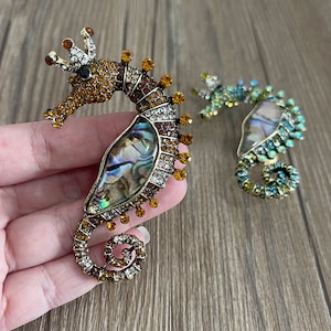 May include: Two seahorse brooches. The larger brooch has a gold-tone metal frame, amber and clear crystals, and an abalone shell inlay. The smaller brooch has a similar design with green and blue crystals. The brooches are decorative accessories.