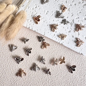 May include: An assortment of small, metallic bee-shaped embellishments in gold, silver, and bronze finishes. The decorative elements are scattered on a textured, off-white surface, with dried ornamental grass in the upper left corner.
