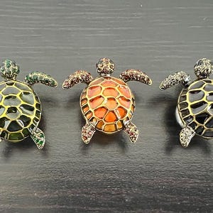 Op de afbeelding: Drie schildpadvormige broches met juwelenaccenten. De schildpadden zijn gemaakt van metaal en hebben een zwarte, groene en oranje emaille afwerking.