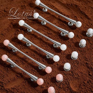 May include: A collection of cabinet hardware, including silver-toned handles with white and marble-patterned spheres, and pink spheres. The handles are arranged on a brown surface. The word "Lotus" is visible in the upper left corner.