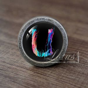 May include: A circular cabinet knob with a silver metal rim. The knob has a black glass centre displaying a colourful, abstract "U" design in pink, blue, and teal. The knob is set against a wooden surface.