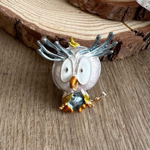 May include: A silver-colored owl brooch with blue and gold accents. The owl has large white eyes, a gold beak, and blue-tipped feathers. The brooch is set on a wooden surface.
