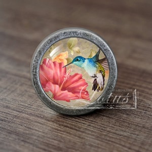 Puede incluir: Un pomo plateado con un inserto de vidrio que presenta un colibrí en vuelo sobre una flor de hibisco rosa.