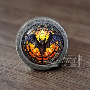 May include: A round, silver-toned cabinet knob with a stained glass design featuring a bat in a circular frame. The stained glass design has shades of orange, yellow, and red.