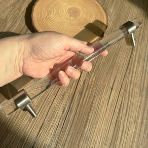 May include: A clear acrylic cabinet pull with brushed nickel fittings. The cylindrical handle is held in a hand, highlighting its transparency and contemporary design. The pull is designed for easy installation on cabinets and drawers.