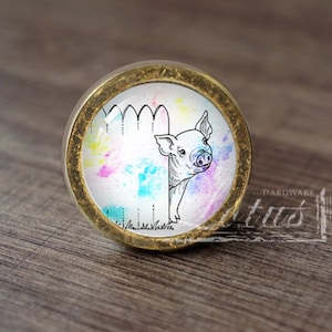 May include: A round, gold-coloured knob with a white background and a colourful watercolour design. A black and white illustration of a pig peeking through a fence is in the centre of the knob.