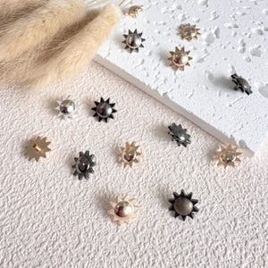 May include: A collection of sun-shaped metal studs in various finishes, including silver, gold, and black. The studs have a starburst design and are scattered on a textured white surface, with some placed on a white rectangular block.