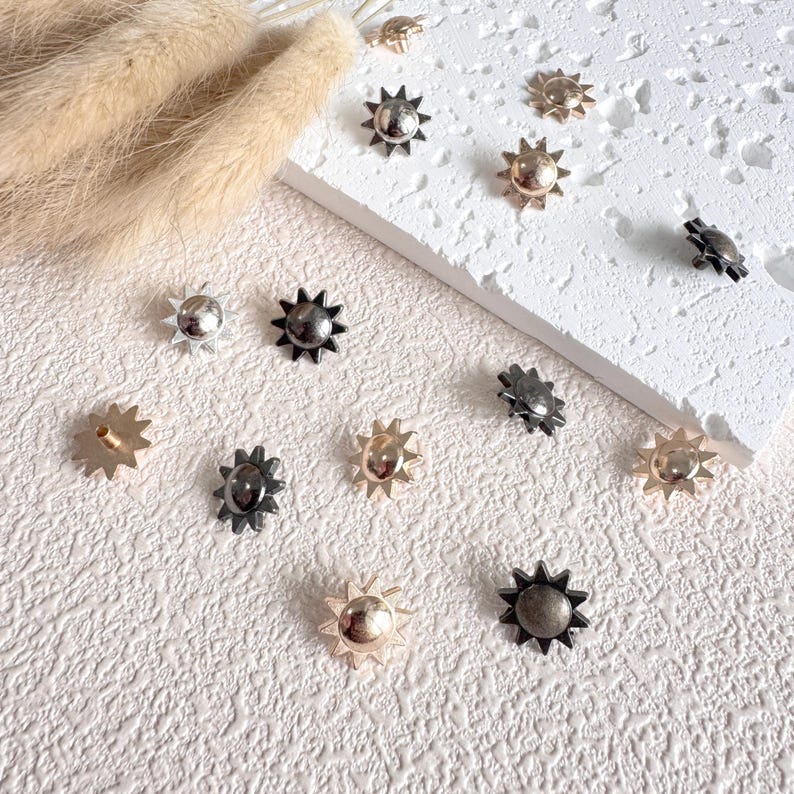 May include: A collection of sun-shaped metal studs in various finishes, including silver, gold, and black. The studs have a starburst design and are scattered on a textured white surface, with some placed on a white rectangular block.