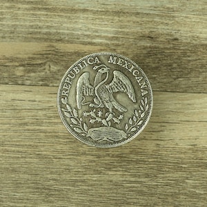 May include: Silver coin with the words "Republica Mexicana" around the edge and a stylised eagle with a snake in its talons in the centre.