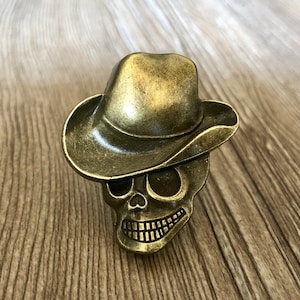 Pode incluir: Uma caveira de metal dourado usando um chapéu de cowboy. A caveira tem um rosto detalhado com órbitas oculares, um nariz e um sorriso com dentes.