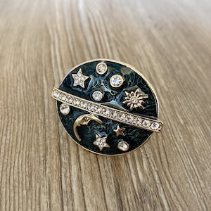 May include: A dark green ring with a gold band and a design of a crescent moon, stars, and a sun with rhinestones. The ring is on a wooden surface.