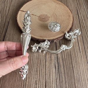 以下が含まれることがあります： シルバーカラーの装飾用金具セット。ハンドル、ノブ、プルが含まれています。これらの部品は、華やかな花柄とスクロールワークのデザインが特徴です。 金具は木製の表面に表示されています。