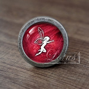 May include: A silver knob with a red background and a white fairy silhouette.