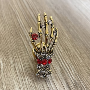 K&ouml;nnte beinhalten: Ein goldener Skeletthandring mit roten und klaren Strasssteinen. Die Hand h&auml;lt eine Schleife mit roten Strasssteinen.