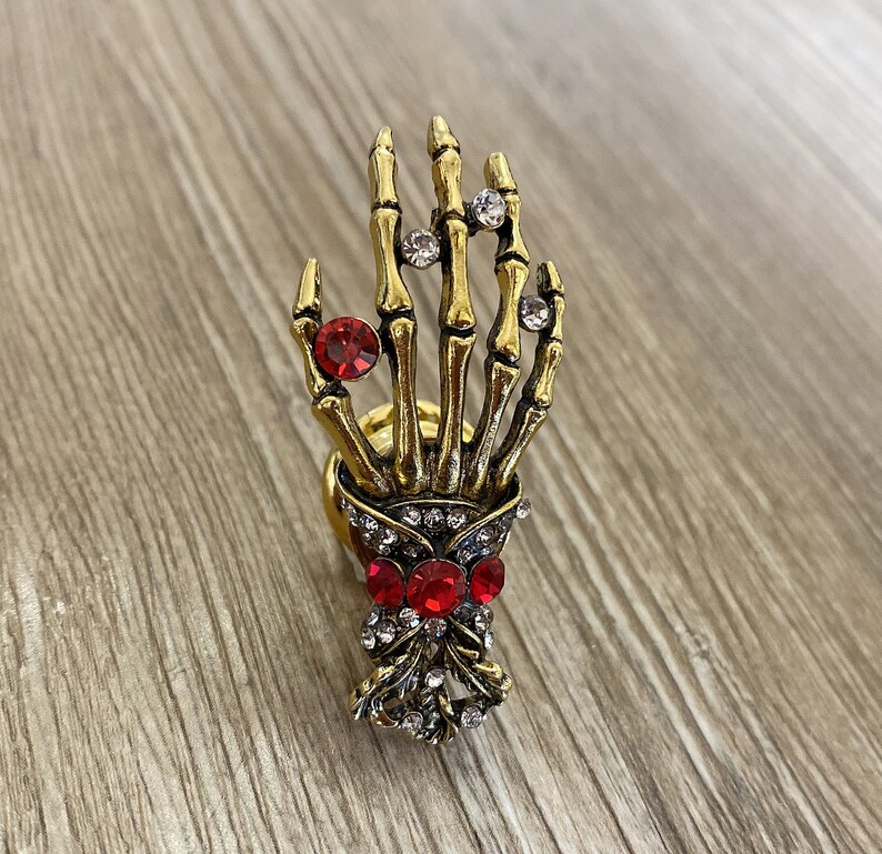 K&ouml;nnte beinhalten: Ein goldener Skeletthandring mit roten und klaren Strasssteinen. Die Hand h&auml;lt eine Schleife mit roten Strasssteinen.