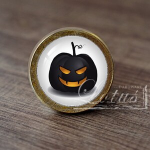 Pode incluir: Um botão de armário redondo de cor latão com um fundo branco e uma abóbora preta com uma cara laranja.