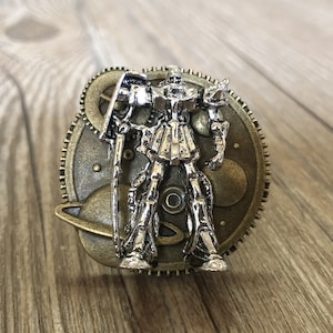 Może przedstawiać: Srebrna figurka robota w stylu steampunk, stojąca na okrągłym, mosiężnym zębatce z planetarnym wzorem. Robot trzyma miecz w prawej ręce.