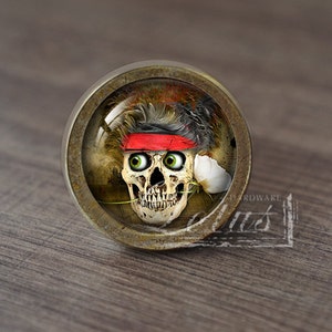 May include: A round, brass-colored cabinet knob with a glass insert featuring a skull with a red bandana and a white flower in its mouth.