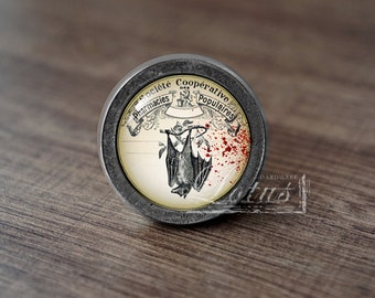 May include: A round, silver-colored cabinet knob with a glass insert featuring a vintage illustration of a bat hanging upside down with blood splatters. The text around the image reads "Societe Cooperative Pharmacies Populaires".