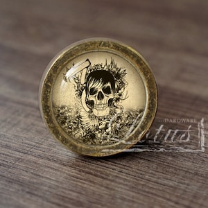 Puede incluir: Un pomo redondo de color latón con un inserto de vidrio que presenta un gráfico de calavera en blanco y negro rodeado de diseños florales.