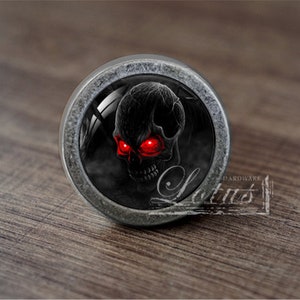 May include: A silver-toned round cabinet knob with a black and red skull design. The skull has red eyes and a black background.