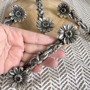 May include: A set of silver-toned metal cabinet pulls featuring a braided handle design with a dimensional daisy flower at each end. The hardware has a metallic finish with dark recessed detailing to highlight the floral and braided textures.