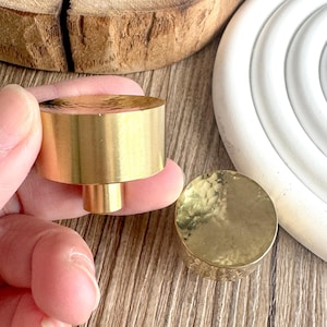 Puede incluir: Dos pomos de latón para armario con textura martillada. Uno se sostiene en una mano, mostrando el cuerpo cilíndrico y el vástago. El otro pomo está plano, mostrando la superficie martillada. Los pomos son de color dorado cálido.