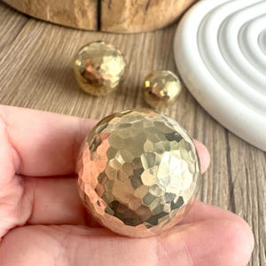 Könnte beinhalten: Nahaufnahme von drei gehämmerten Messingkugeln. Die größte Kugel wird in einer Hand gehalten, mit zwei kleineren Kugeln im Hintergrund. Die Kugeln haben eine strukturierte, gekörnte Oberfläche und eine warme, goldene Farbe. Dekoratives Zubehör.