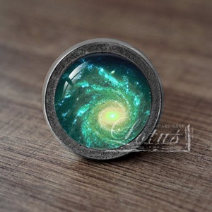 Puede incluir: Un pomo redondo de color plateado con un inserto de vidrio que presenta un diseño de galaxia verde y azul en espiral.