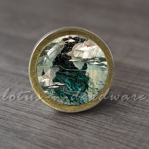 May include: A round, gold-toned cabinet knob with a glass insert featuring a black and white illustration of a sea creature with a teal and blue background.