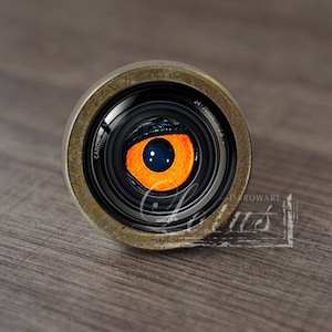 Puede incluir: Primer plano de un objetivo de cámara con un ojo naranja en su interior. El objetivo está hecho de metal y tiene un acabado negro. El ojo está rodeado por un anillo negro y tiene una pupila azul.