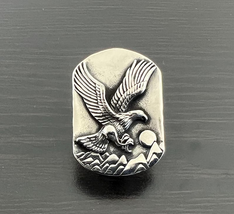 May include: A silver-toned rectangular pendant featuring a detailed relief of an eagle in flight. The eagle is depicted with outstretched wings, soaring above a mountain range and a full moon. The pendant has a slightly aged, antique appearance.