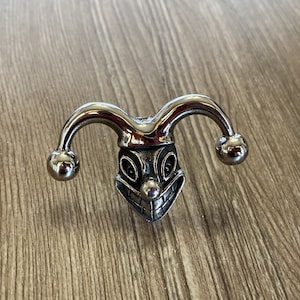 Könnte beinhalten: Ein silberner Jester-Kopfring mit einem breiten, zahnigen Grinsen und einer spitzen Mütze. Der Ring ist aus Metall und hat eine polierte Oberfläche.