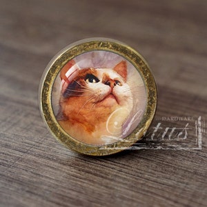 以下が含まれることがあります： 猫が上を見ている絵が描かれたガラス製のインサートが付いた、丸い金色調のキャビネットノブ。猫は白い毛にオレンジと茶色の模様があります。