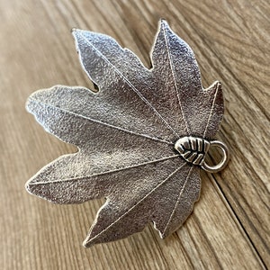 Pode incluir: Um amuleto de folha de bordo de metal prateado com uma superf&iacute;cie texturizada e um pequeno la&ccedil;o na parte de tr&aacute;s para prender a joias ou outros artesanatos.