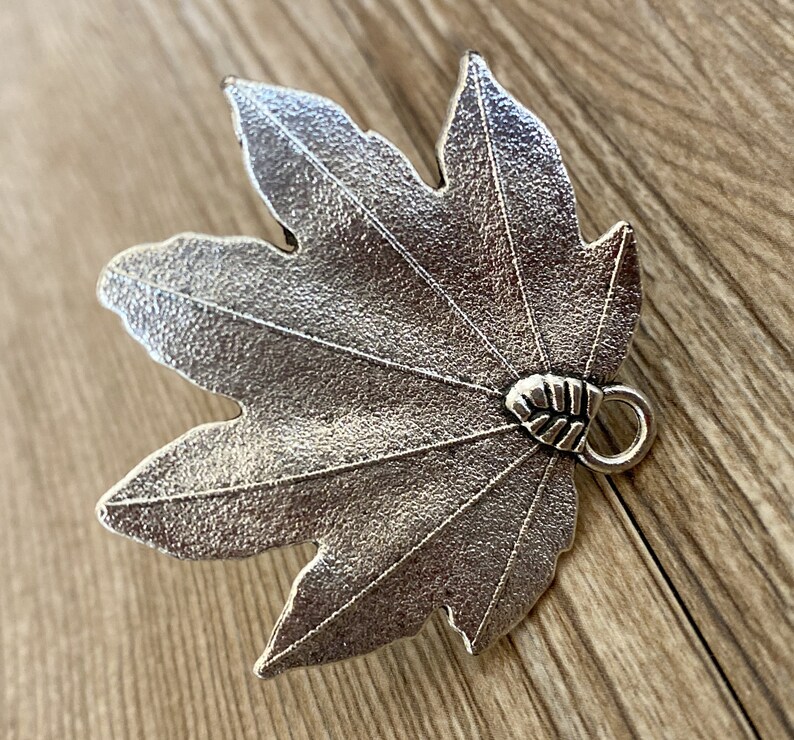 Pode incluir: Um amuleto de folha de bordo de metal prateado com uma superf&iacute;cie texturizada e um pequeno la&ccedil;o na parte de tr&aacute;s para prender a joias ou outros artesanatos.