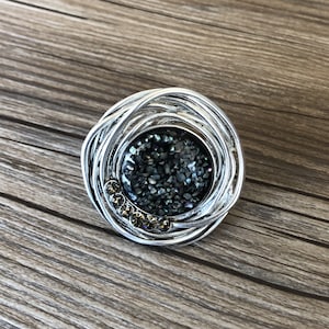 May include: A silver ring featuring a black and iridescent stone set within a circular wire design. The stone is surrounded by small clear rhinestones.