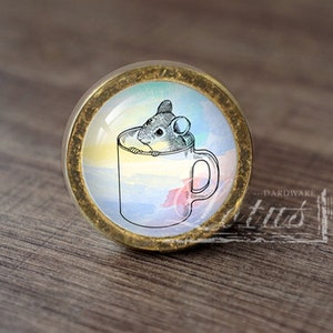 May include: A round, brass-colored cabinet knob with a glass insert featuring a watercolor illustration of a small mouse peeking out of a teacup. The teacup is white with a blue and pink watercolor design.
