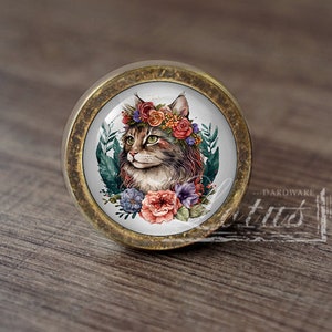 Könnte beinhalten: Ein runder, messingfarbener Schrank-Knopf mit einem weißen Keramik-Einsatz, der eine Katze mit einer Blumenkrone zeigt. Die Katze hat braunes und weißes Fell und grüne Augen. Die Blumen sind rosa, rot und lila.