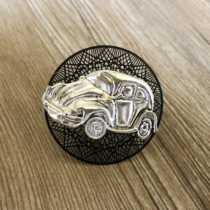 Op de afbeelding: Een zilverkleurige autohanger met een zwarte ronde achtergrond. De autohanger is een gedetailleerde weergave van een klassieke auto.