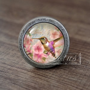 Puede incluir: Un pomo plateado con un inserto de vidrio que presenta un colibrí en vuelo sobre flores rosas.