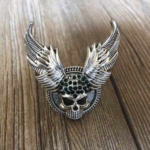 Könnte beinhalten: Silberner Totenkopf mit grünen Strasssteinen und Flügeln.