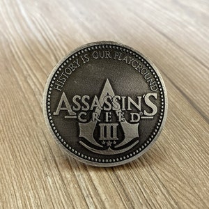 May include: A silver-colored coin with the words "History is our playground" and "Assassin's Creed III" around the edge. The center of the coin features a stylized assassin's symbol.